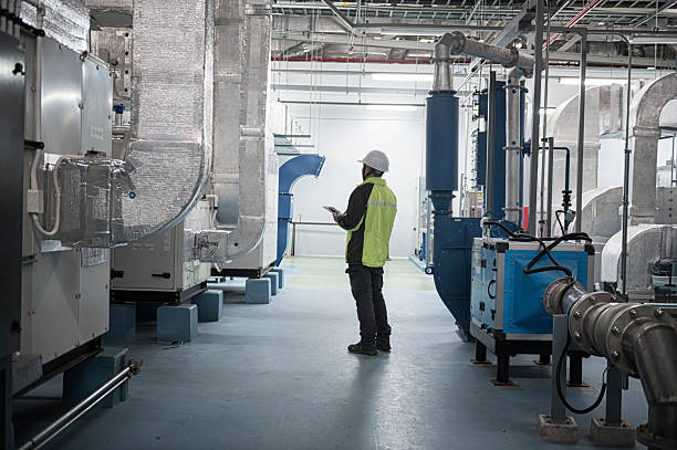 professional Asian engineer or technician in safety gear performs a routine inspection using a digital tablet inside a modern industrial facility. Surrounded by high-tech piping and equipment, he checks operational systems in a controlled, clean, and safe work environment.